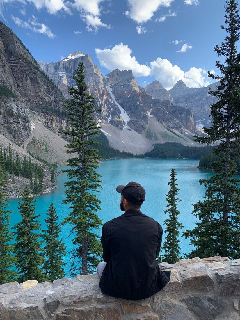 A person sitting on a stone wall looking out over a turquoise lake ringed by mountains